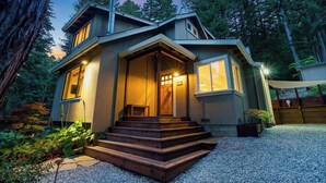 A modern house with a wooden staircase leading to the entrance, surrounded by a gravel driveway and a garden with various plants. - Redwood Oasis - Luxe Santa Cruz Cabin w/ Hot Tub (Ben Lomond)