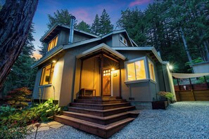 A modern house with a wooden staircase leading to the entrance, surrounded by a gravel driveway and a garden with various plants. - Redwood Oasis - Luxe Santa Cruz Cabin w/ Hot Tub (Ben Lomond)