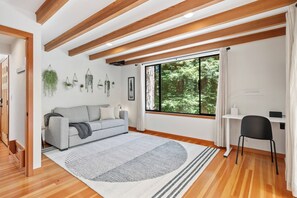 Una sala de estar moderna con un sofá gris, un tapete redondo, vigas de madera, un ventanal con vistas a la vegetación y un escritorio pequeño con una silla.