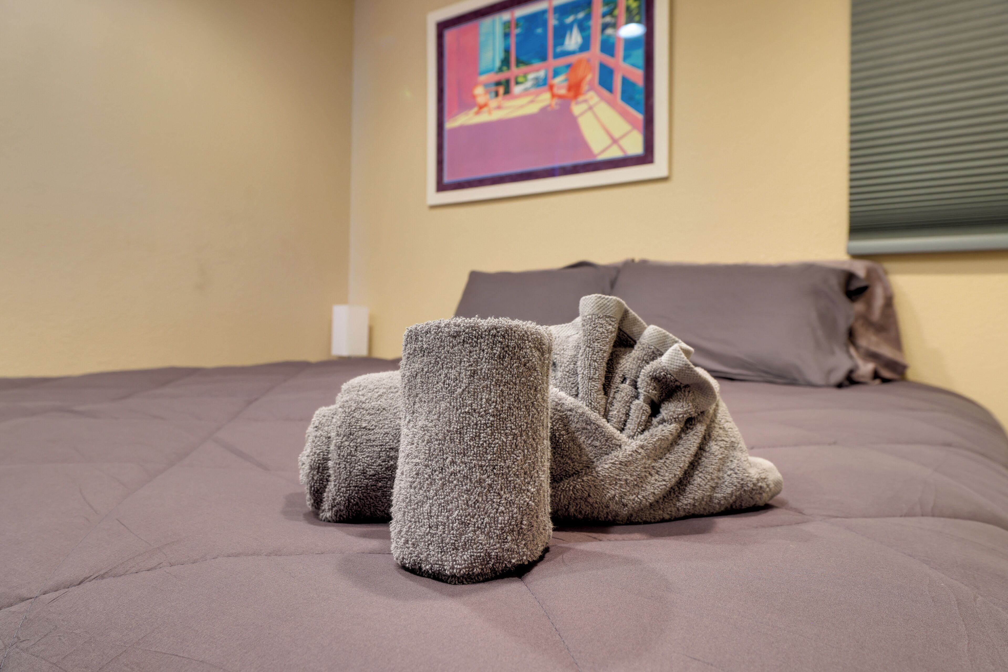 Ein Bett mit grauer Decke und Kissen, ein Bild an der Wand und ein Fenster mit Jalousien.