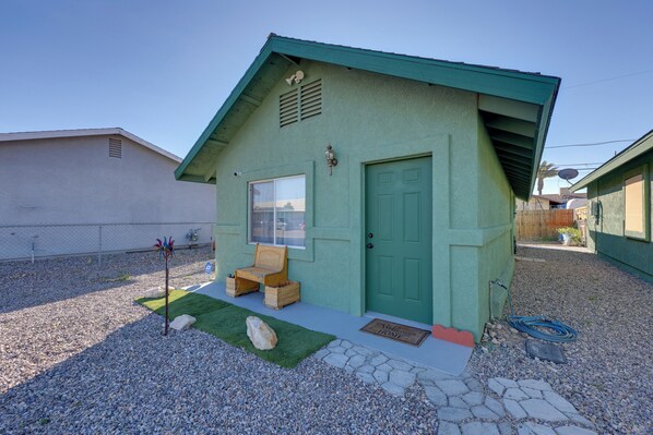 A small, single-story house with a green door and a window, a small porch with a bench, and a gravel driveway. - Near Parks, Shops & Eats! Bullhead City Casita (Bullhead City)