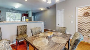 Un comedor con una mesa de madera, sillas estampadas y una cocina al fondo.