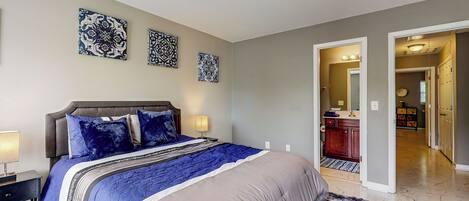 Een slaapkamer met een bed, nachtkastjes en decoratie aan de muur. Er is een gang naar een badkamer en een keuken.