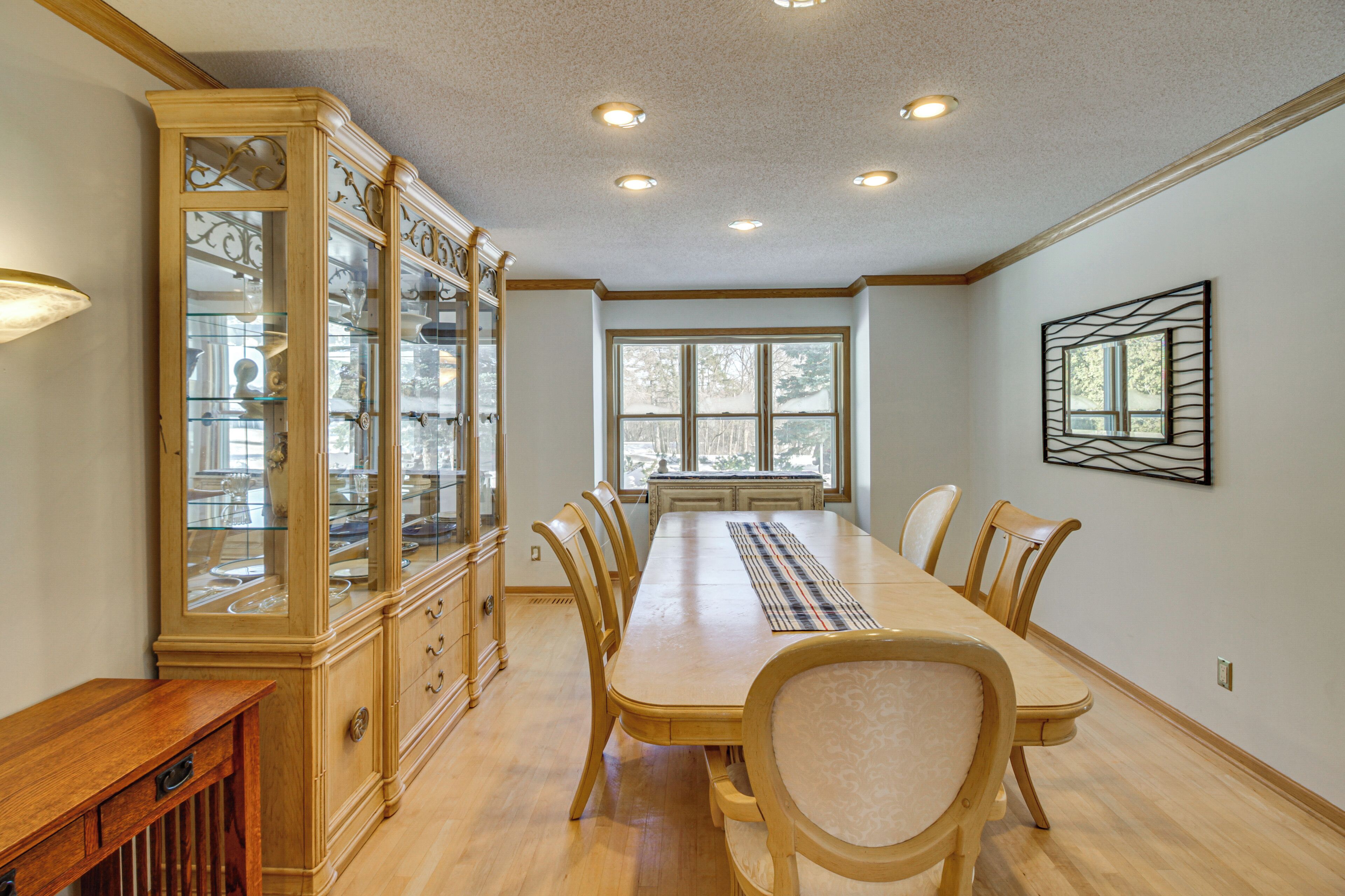A dining room with a long wooden table, six chairs, a glass-fronted cabinet, and a window with a view of trees.