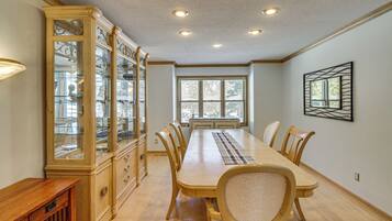 A dining room with a long wooden table, six chairs, a glass-fronted cabinet, and a window with a view of trees.