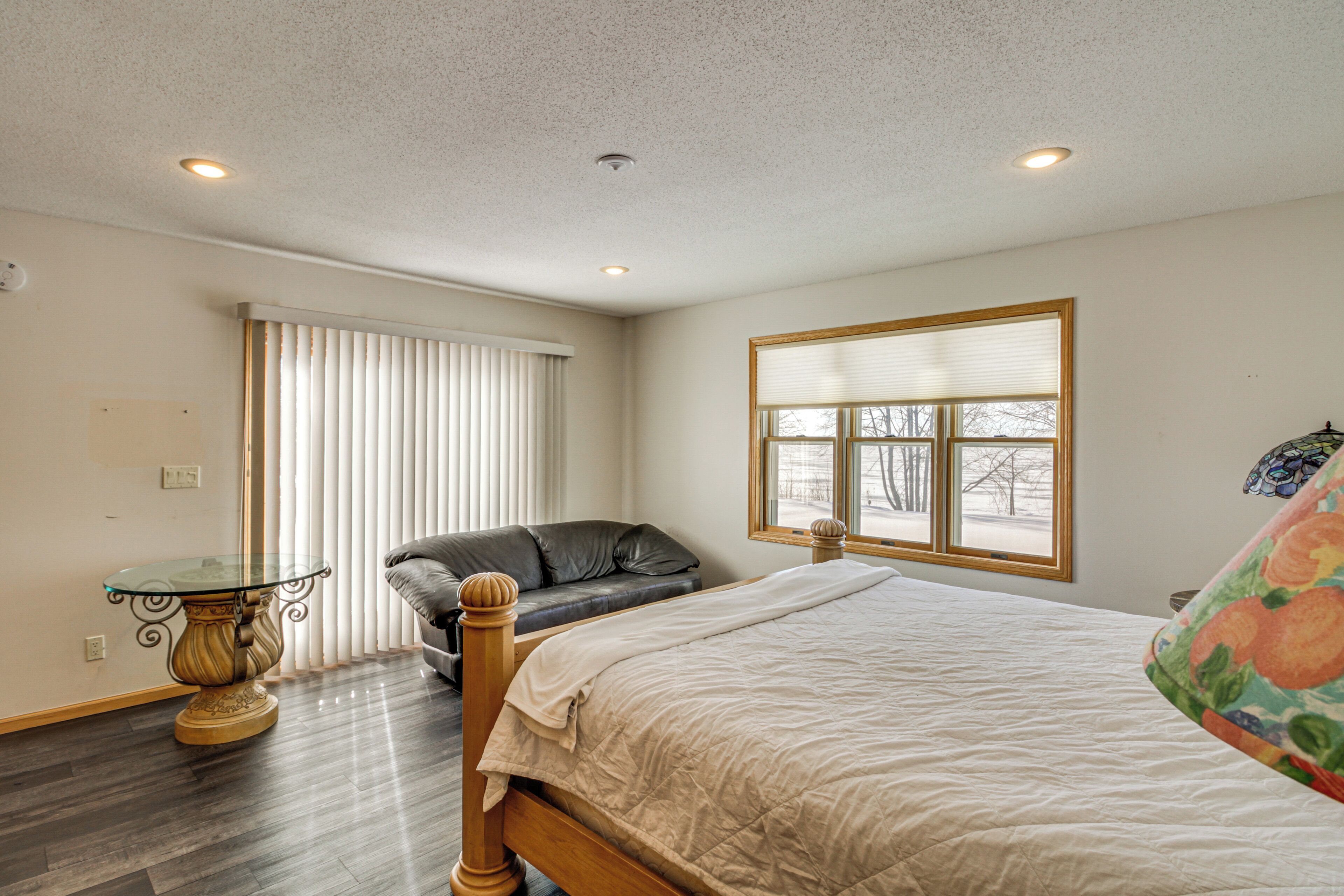 A bedroom with a bed, a sofa, a glass-top table, and a window with blinds.