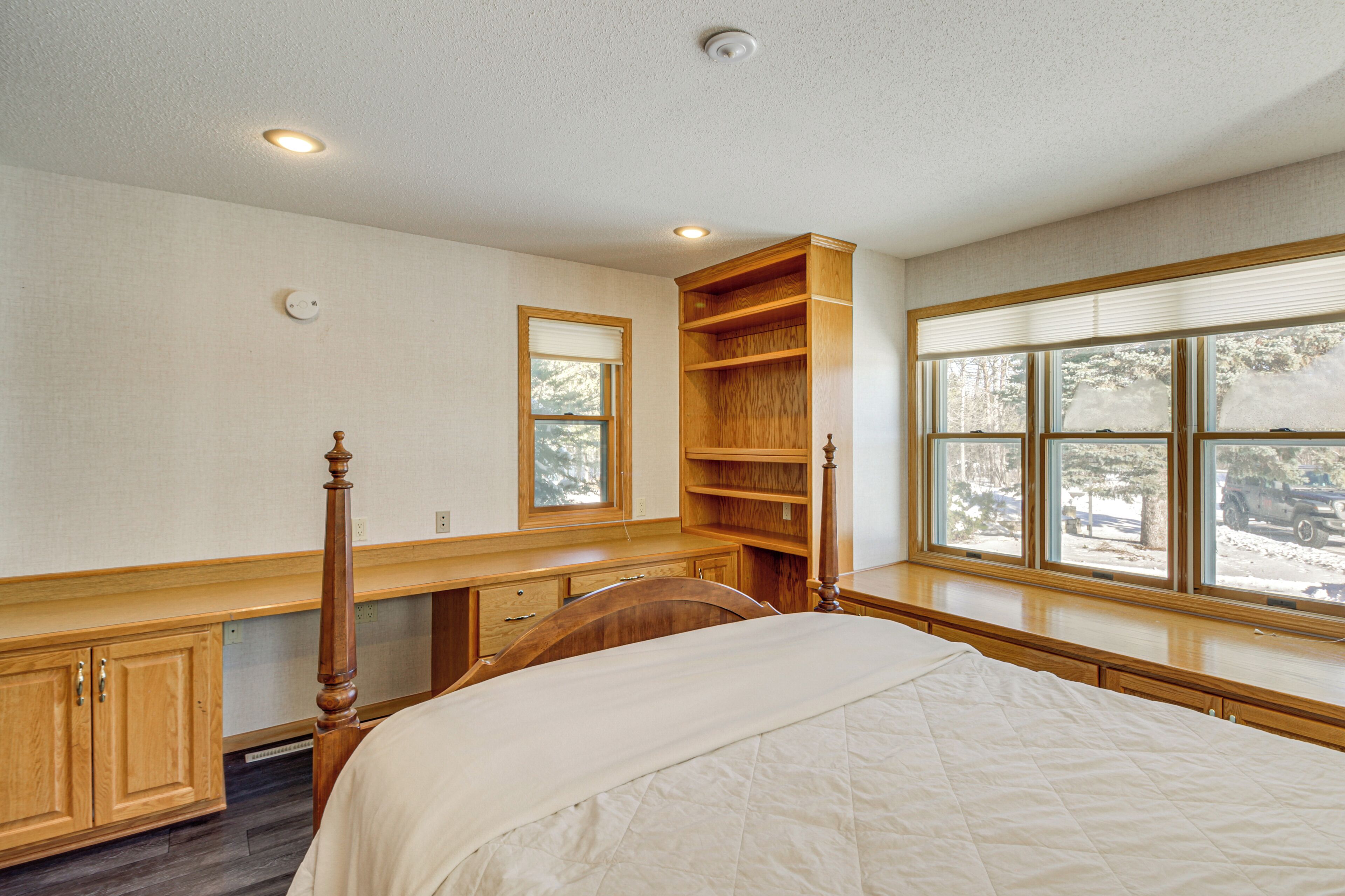 A bedroom with a wooden bed, a nightstand, a built-in shelf, a window with blinds, and a ceiling light.