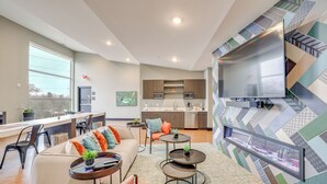 Un salon moderne avec un canapé, une table basse et des fauteuils. On aperçoit, au fond, une cuisine avec une grande fenêtre et un coin repas.