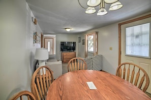 Une salle à manger avec une table et des chaises en bois, un téléviseur en arrière-plan et une fenêtre avec des stores.
