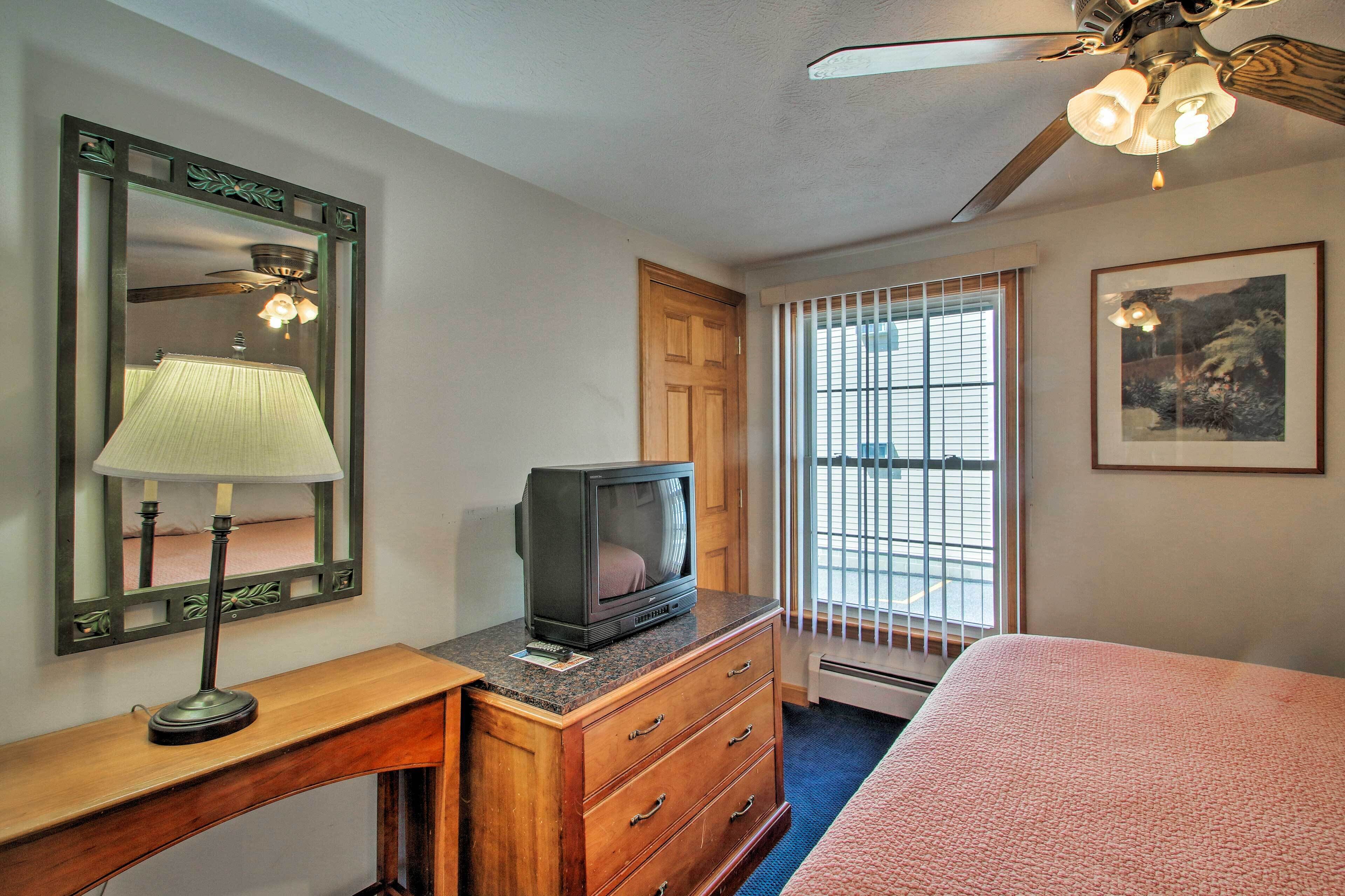 Ein Hotelzimmer mit einem hölzernen Schminktisch, einem Fernseher darauf, einem Spiegel mit dekorativem Rahmen, einem Deckenventilator und einem Fenster mit Jalousien.