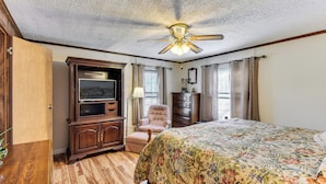 A bedroom with a wooden floor, a bed with a floral bedspread, a ceiling fan, a television on a wooden cabinet, a chair, and a dresser. - Countryside Haven w/ Spacious Yard in Tamassee! (Tamassee)