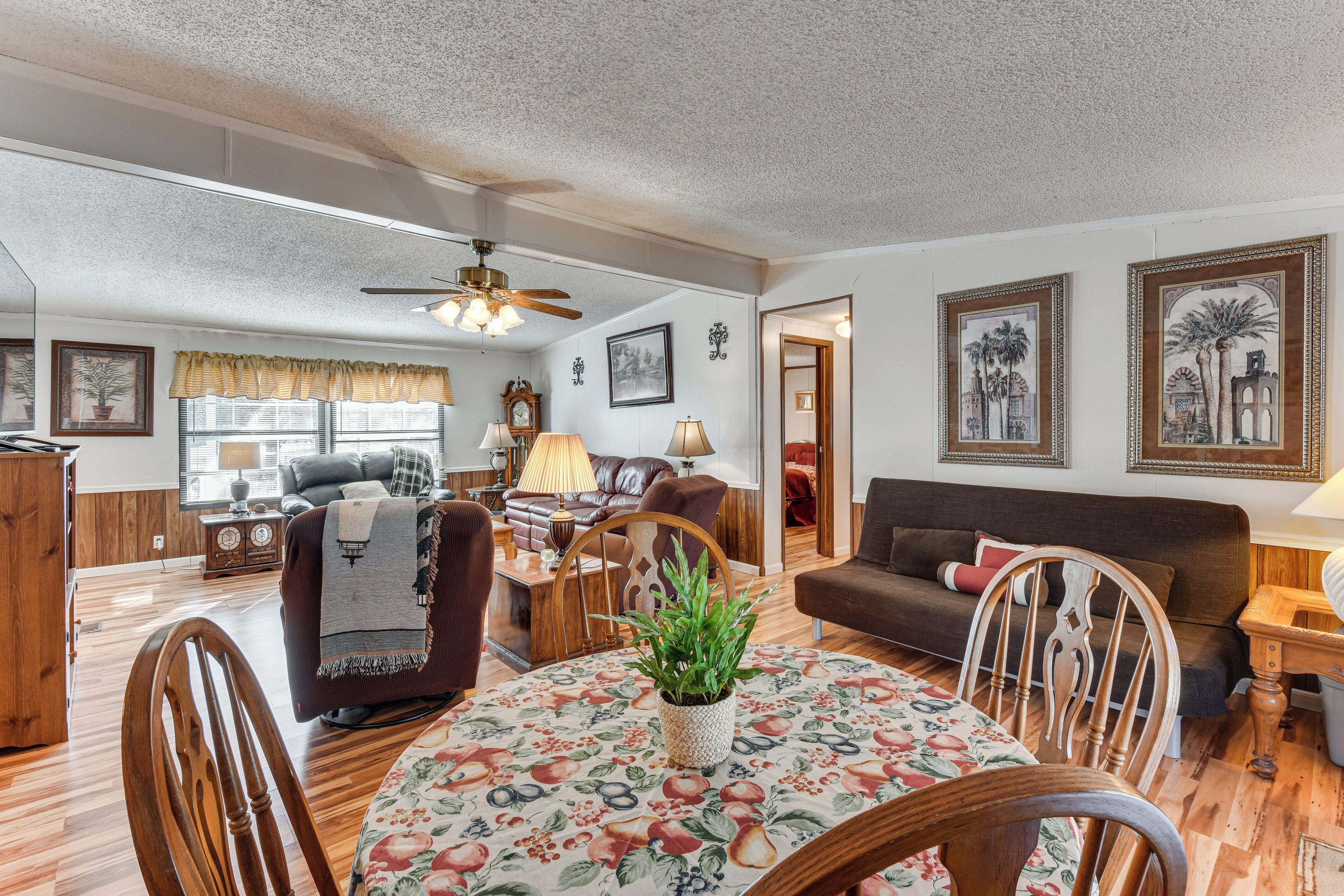 A living room with a wooden floor, a round table with a floral tablecloth, a sofa, a chair, and a ceiling fan.