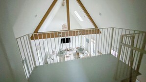 Magas mennyezetű szoba, fehér mennyezettel és fa gerendákkal, fehér korláttal, valamint asztallal és székekkel.