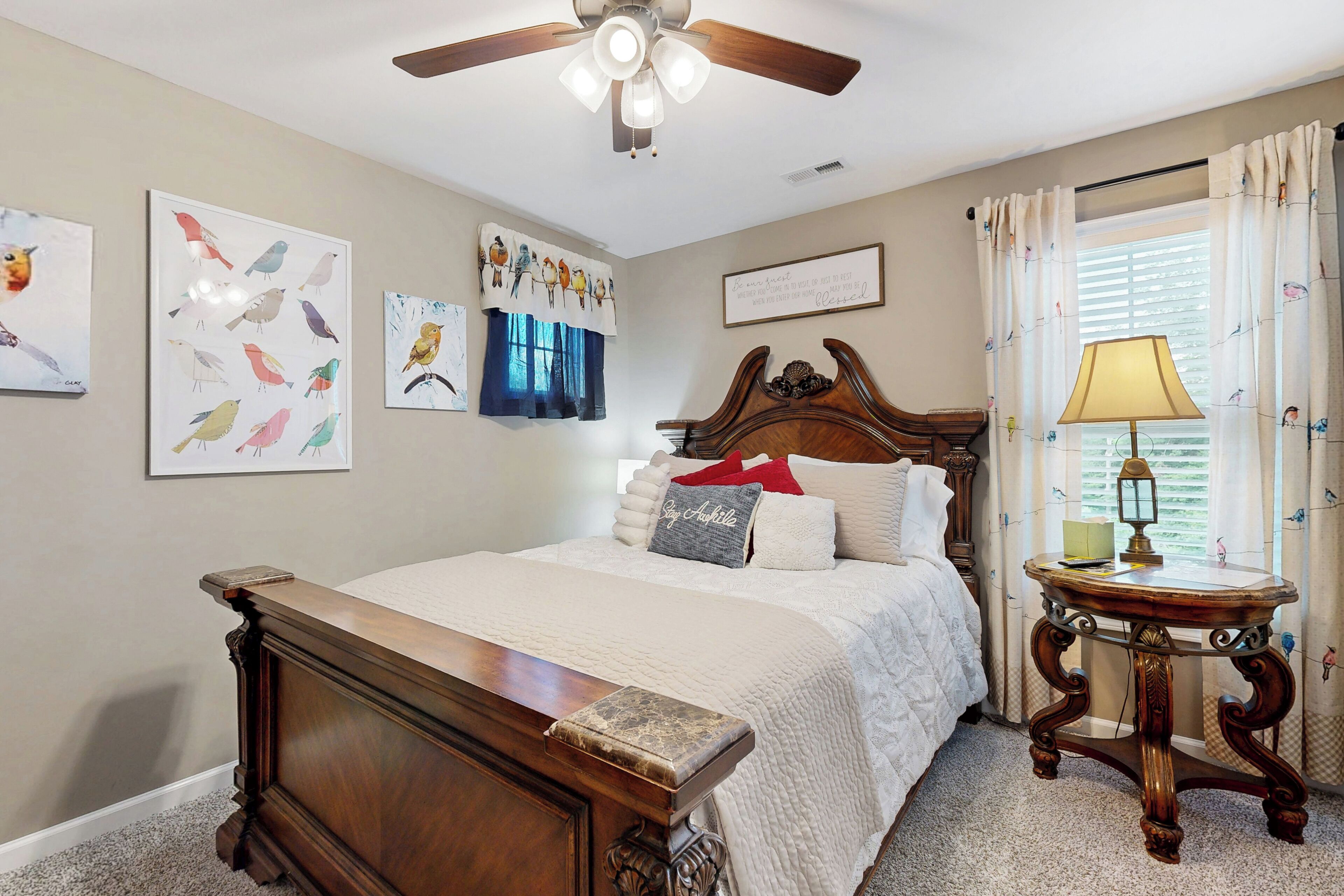 A bedroom with a wooden bed, a ceiling fan, wall art, a bedside table with a lamp, and a window with curtains.