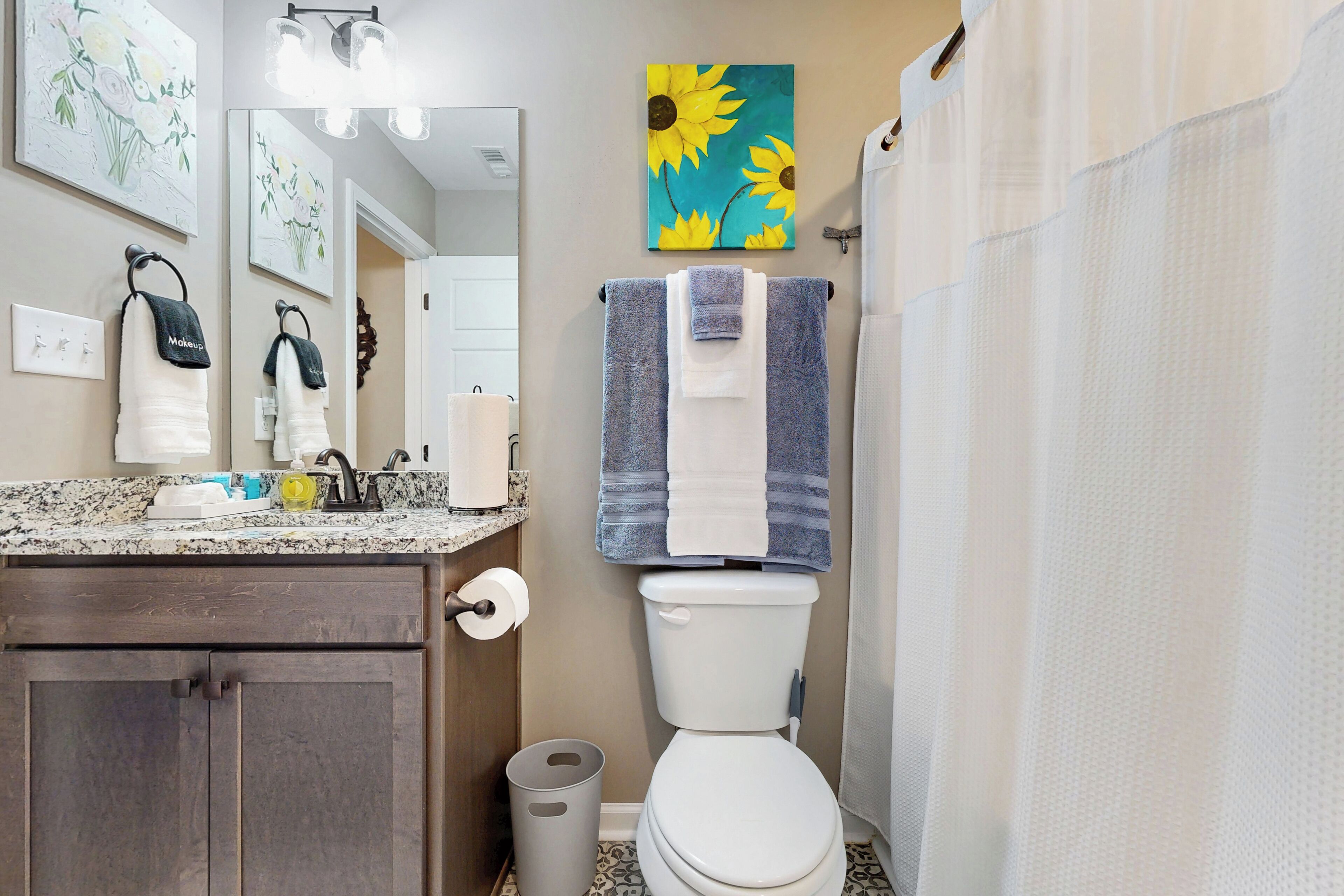 A bathroom with a toilet, sink, and a mirror. There are towels hanging on the wall and a trash can next to the toilet.