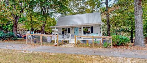 A small, single-story house with a porch, surrounded by trees and a fenced yard.