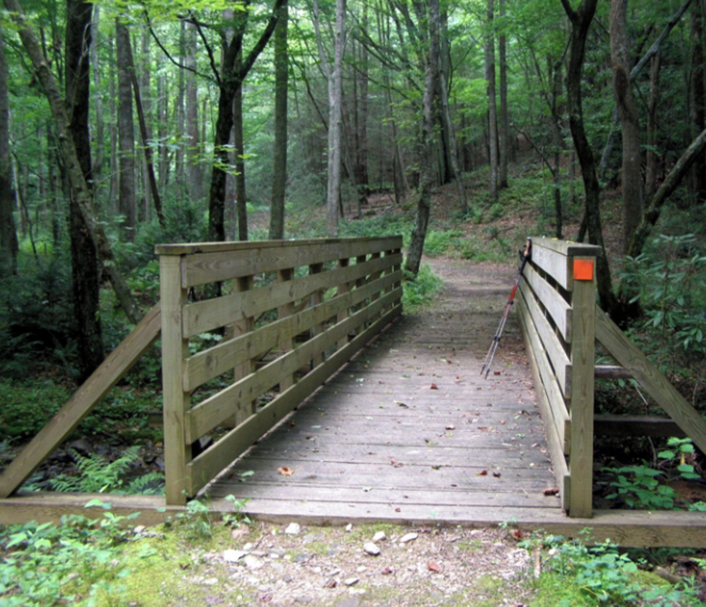 Un pont en bois, situé dans une zone boisée, avec une rambarde en métal et un panneau rouge sur le côté droit.