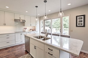 A modern kitchen with white cabinets, a large island with a sink, pendant lights, and a view of the outdoors through large windows. - Plymouth House: Walk to Beach & Medicine Lake! (Plymouth)