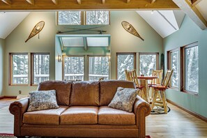 A spacious room with a leather sofa, wooden dining table, and chairs. Large windows provide views of trees. - Private Peninsula! Laurel Lake Retreat w/ 3 Piers (Three Lakes)