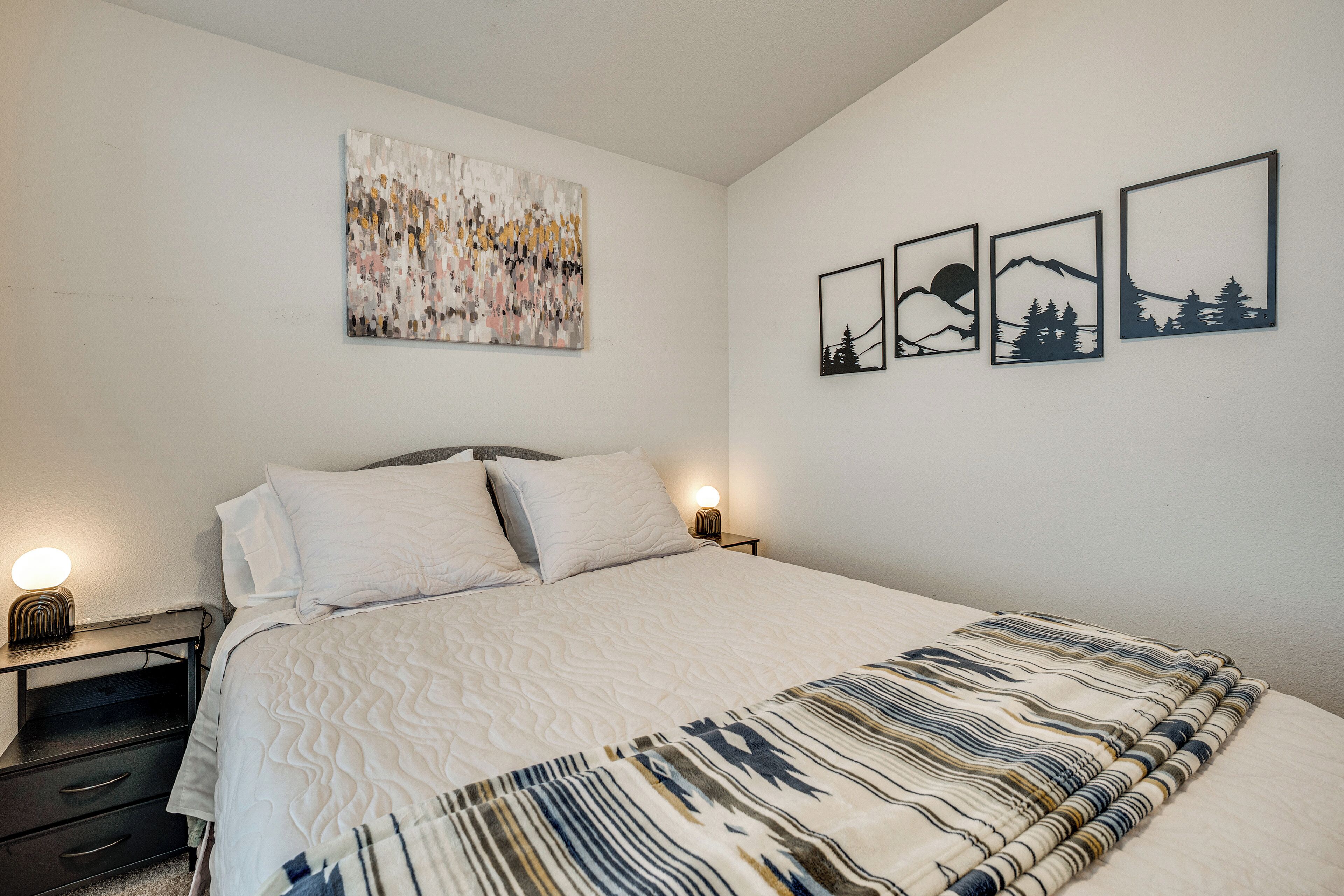 Una camera da letto con un letto, un comodino, un quadro astratto appeso al muro e tre stampe raffiguranti paesaggi.