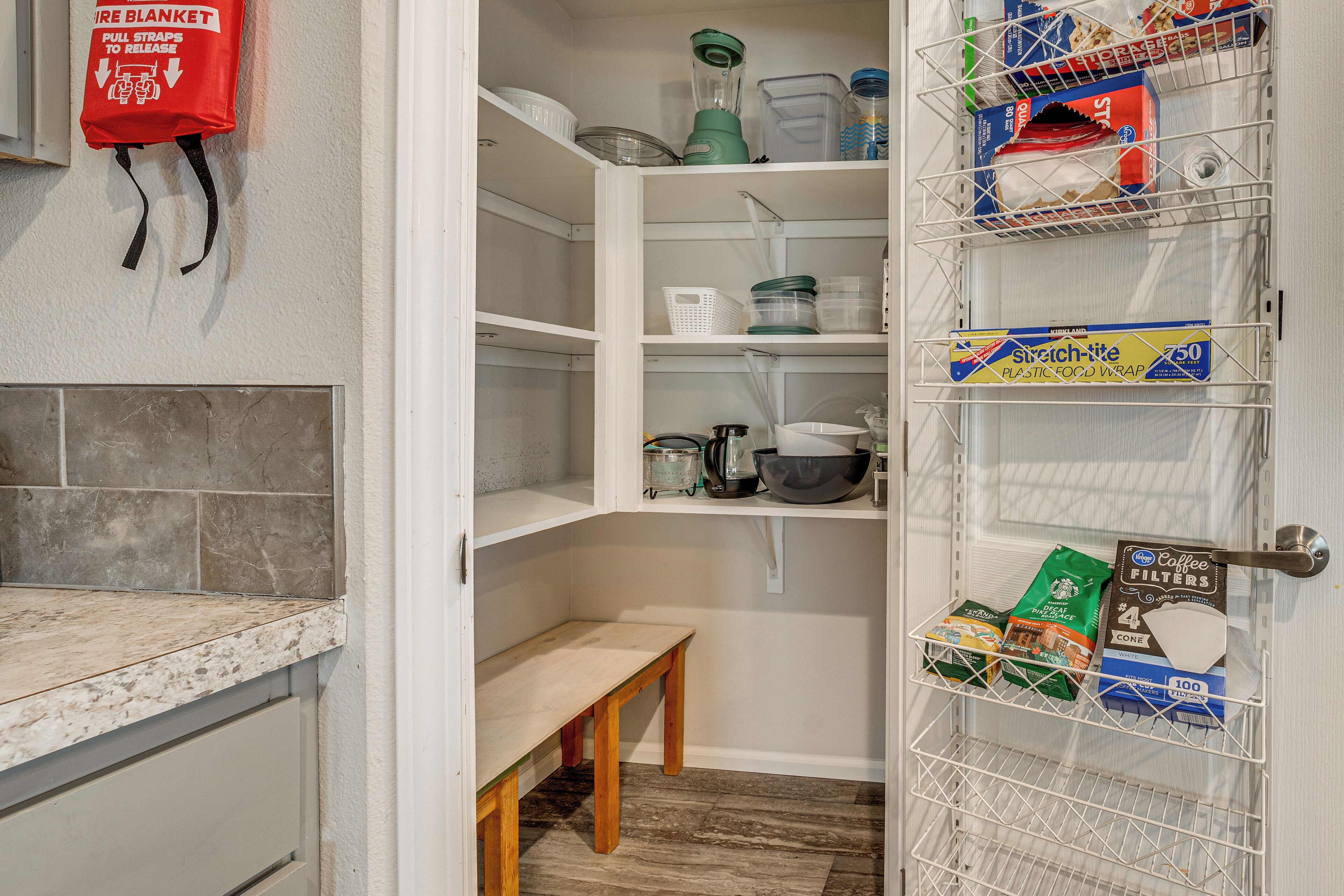 A kitchen with a countertop, a wooden bench, and a pantry with shelves containing kitchen items and storage containers.
