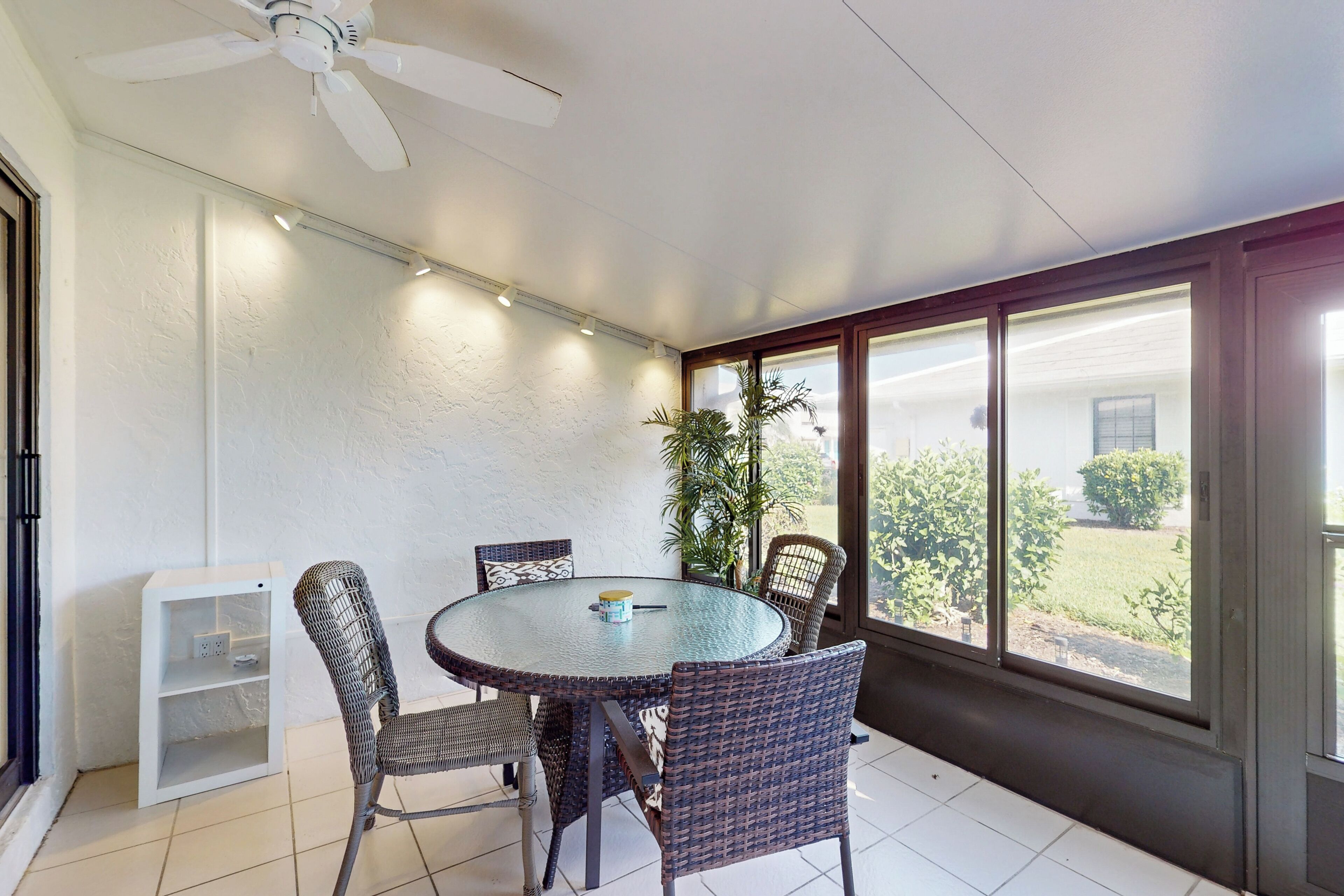 Una veranda riparata con un tavolo rotondo in vetro, sedie in vimini, una ventola a soffitto e vista sull'esterno.