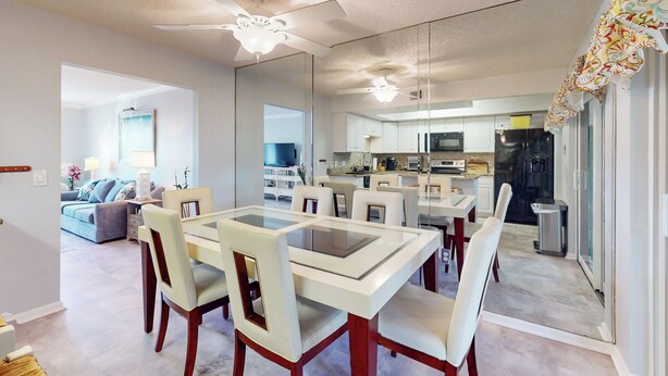Un comedor moderno con una mesa de vidrio y sillas de madera, conectado a una cocina con gabinetes blancos y un ventilador de techo.