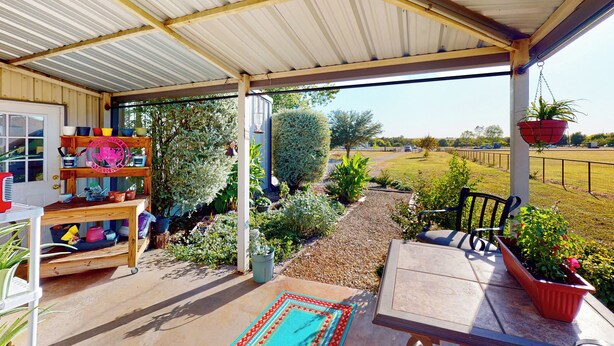 A covered patio area with a wooden table and chairs, surrounded by potted plants and a garden.
