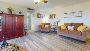A cozy living room with a wooden floor, a ceiling fan, a wooden cabinet, two armchairs, a sofa, and a small table with a lamp.