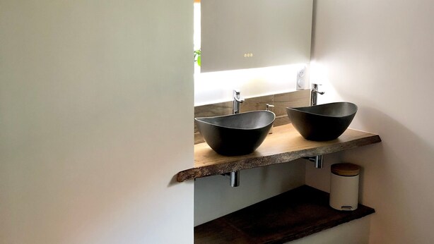 Un baño con dos lavamanos negros, un estante de madera, un cubo de basura blanco y una cesta tejida.