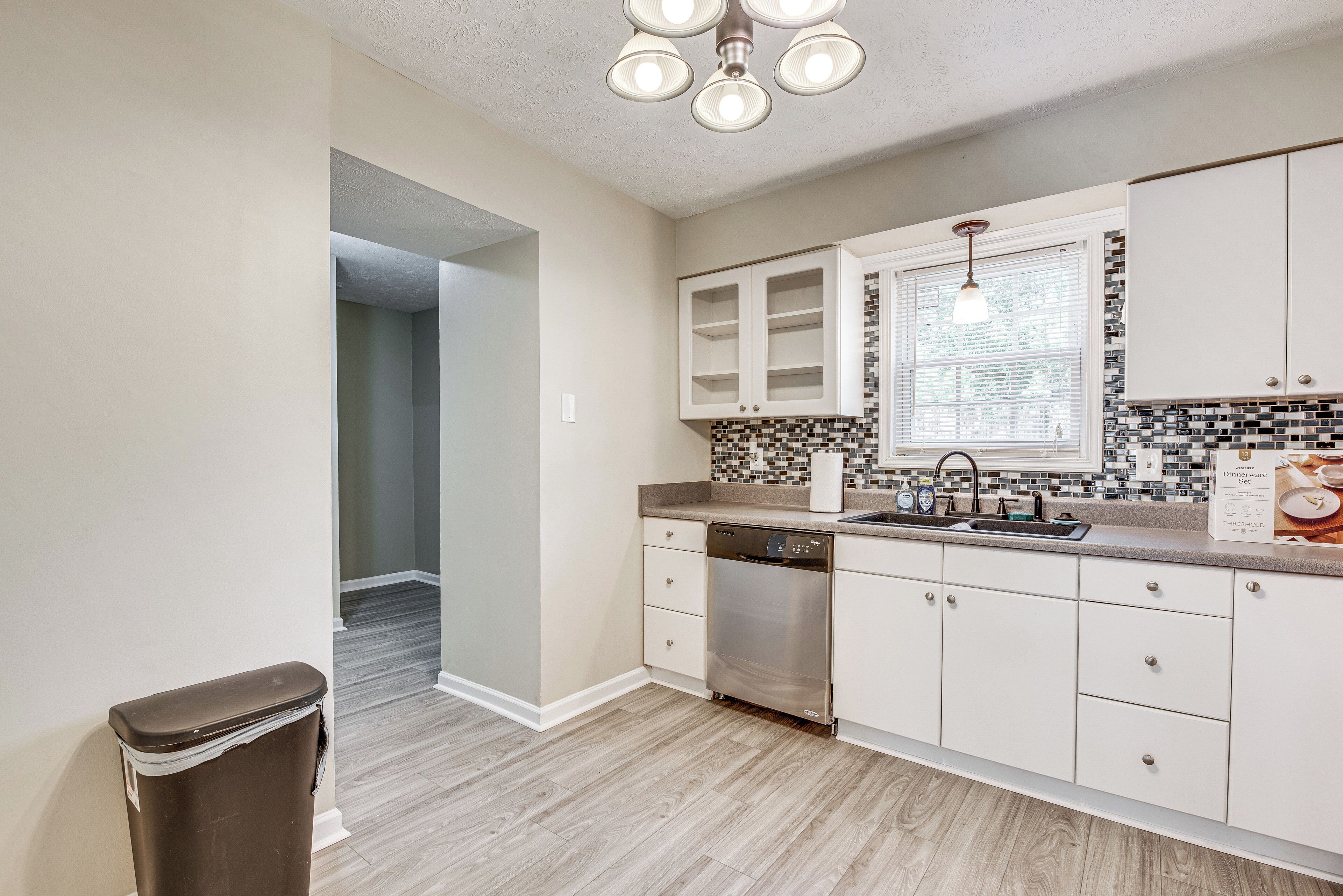 A kitchen with white cabinets, a dishwasher, a window with blinds, and a tiled backsplash.