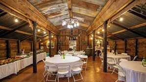 Un espace de restauration de style rustique, aménagé dans une ancienne grange, avec des tables rondes et des chaises blanches, des poutres en bois et un lustre rustique.