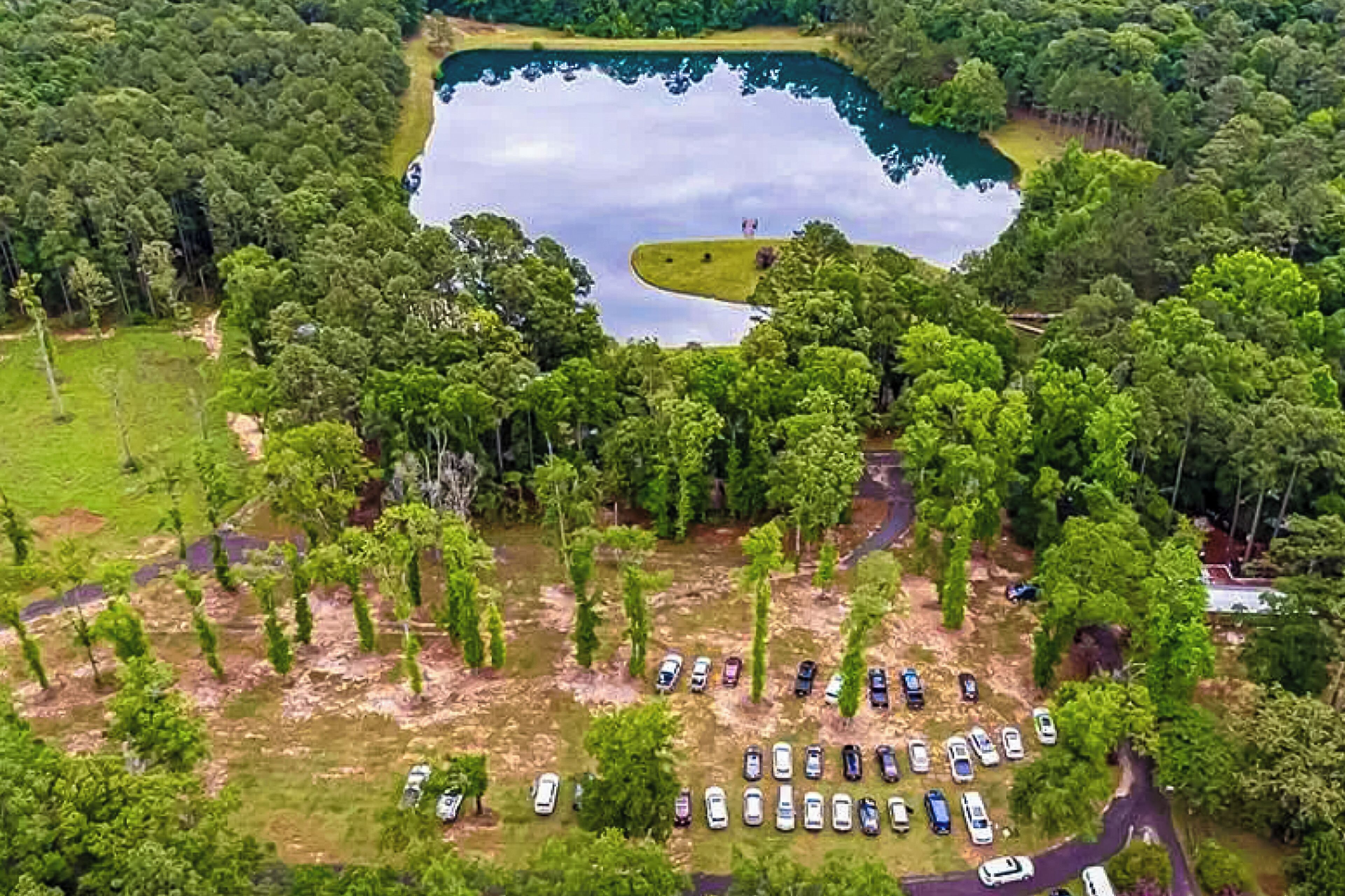 An aerial view of a campground with a large lake, surrounded by dense trees and a parking area with multiple cars.
