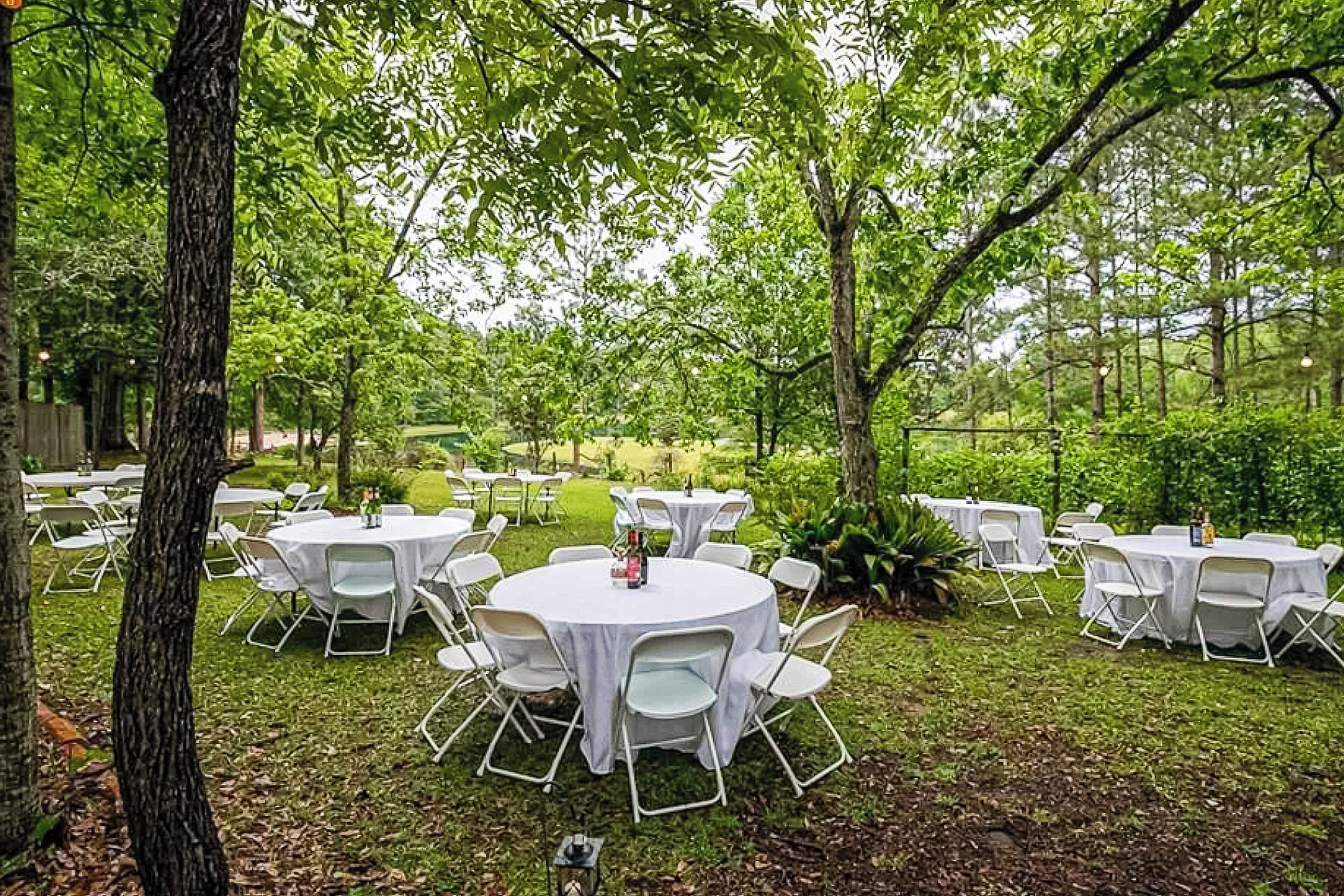 An outdoor venue with round tables and white chairs arranged on a grassy area, surrounded by trees.