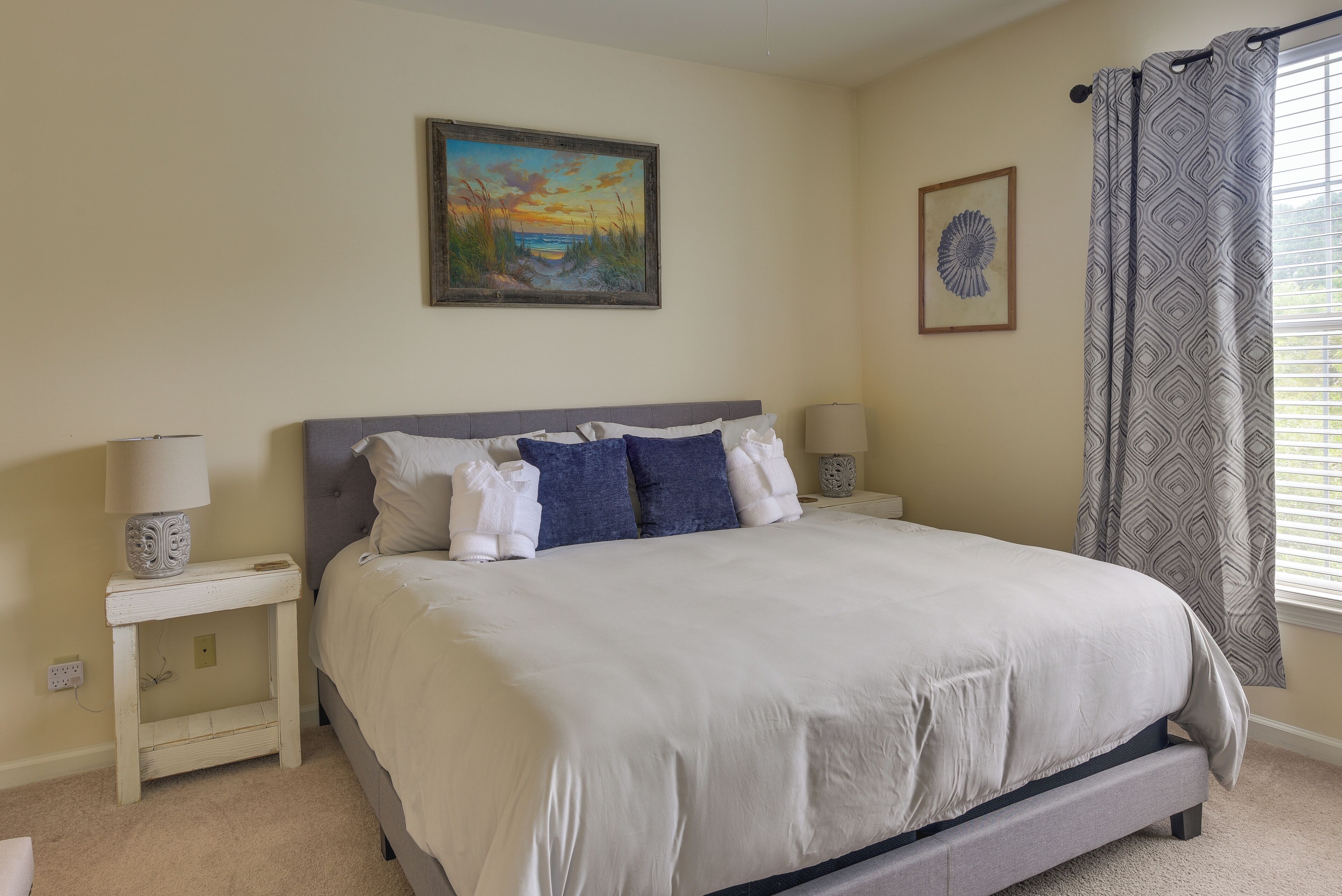 A neatly made bed with a grey headboard, two bedside tables with lamps, and a painting above the bed.