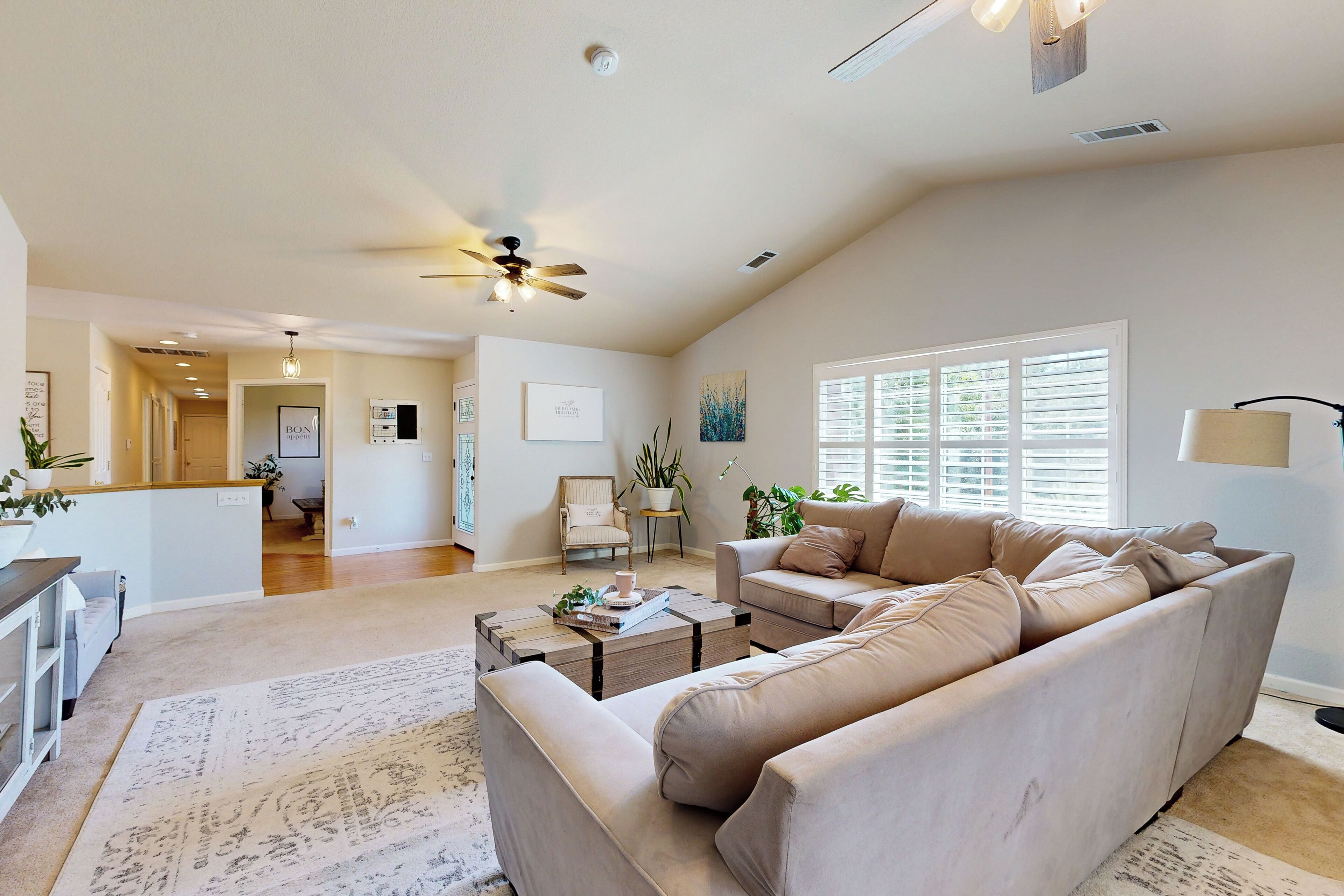 A spacious living room with a large beige sectional sofa, a coffee table, and a dining area with a kitchen in the background.