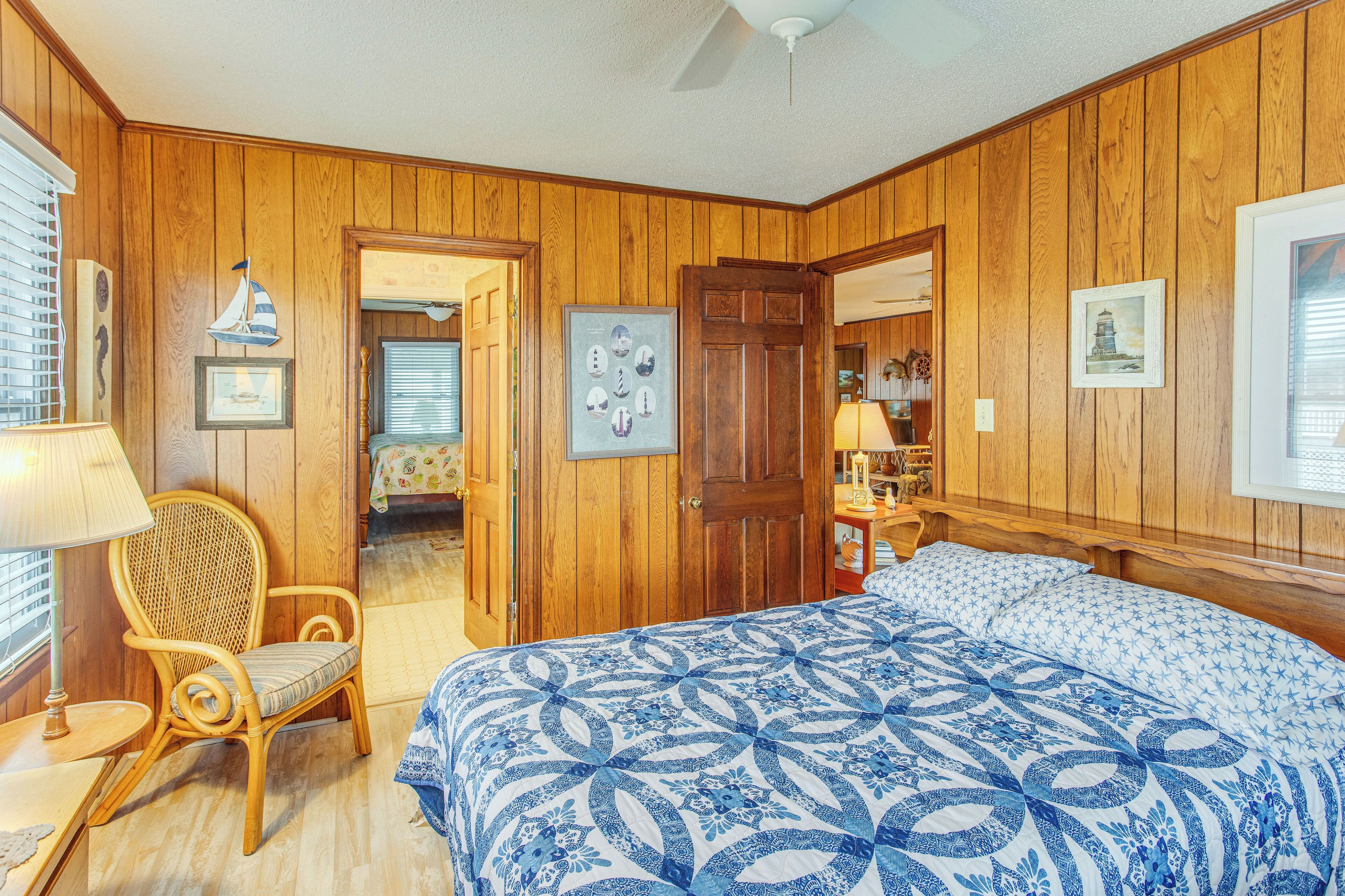 A bedroom with wooden paneling, a bed with a blue and white patterned bedspread, a wicker chair, and a door leading to another room.