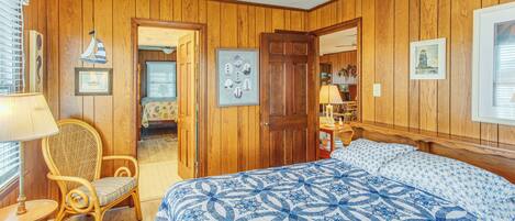A bedroom with wooden paneling, a bed with a blue and white patterned bedspread, a wicker chair, and a door leading to another room.
