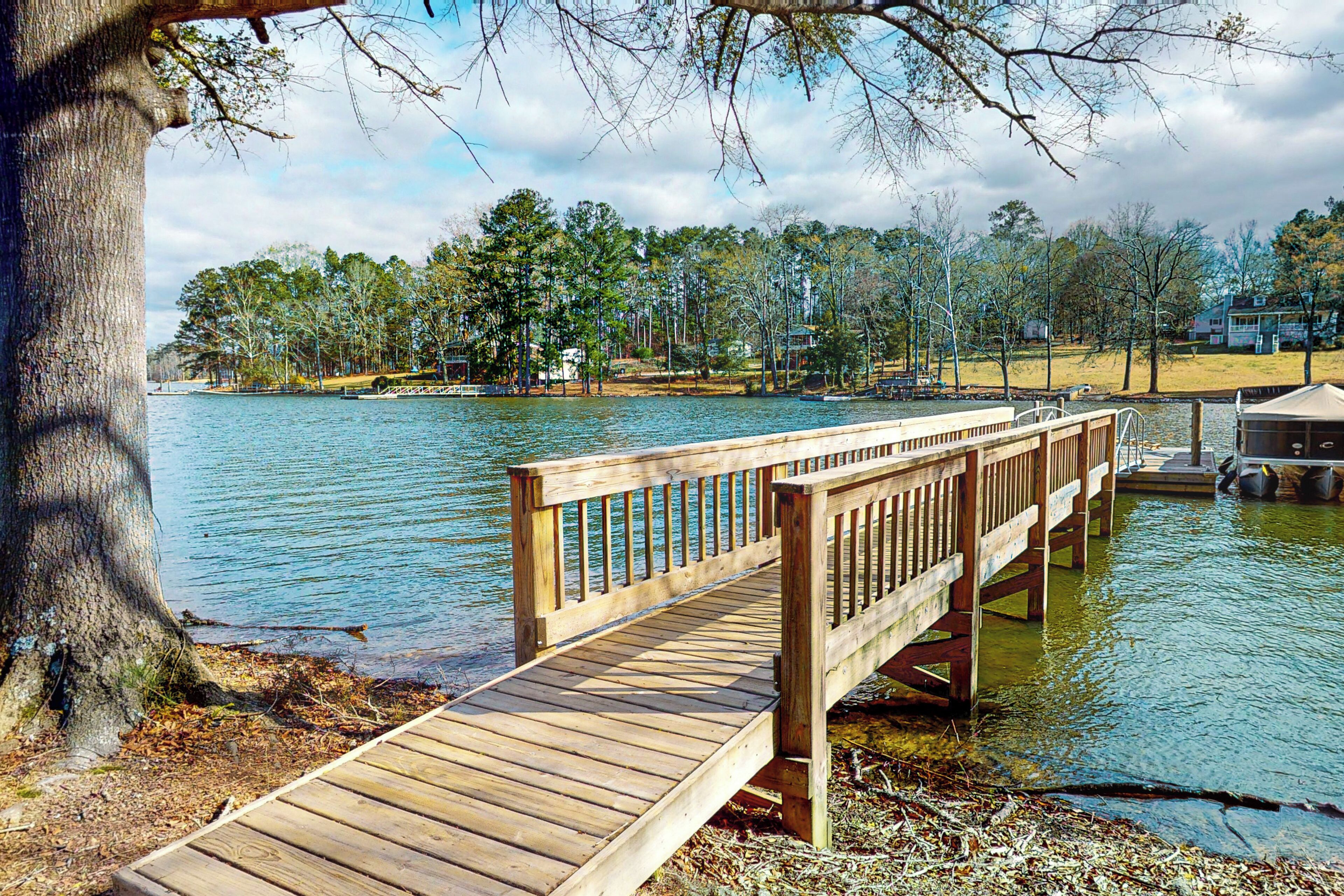A wooden dock extending over calm water, surrounded by trees and a distant shoreline with buildings.