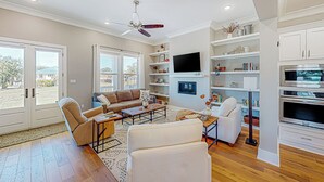 A spacious living room with a large sofa, a coffee table, and a TV mounted on the wall. There are shelves with decorative items and a ceiling fan. - 3BR Home Near Beach w/ Views, Deck & Grill (Gulfport)