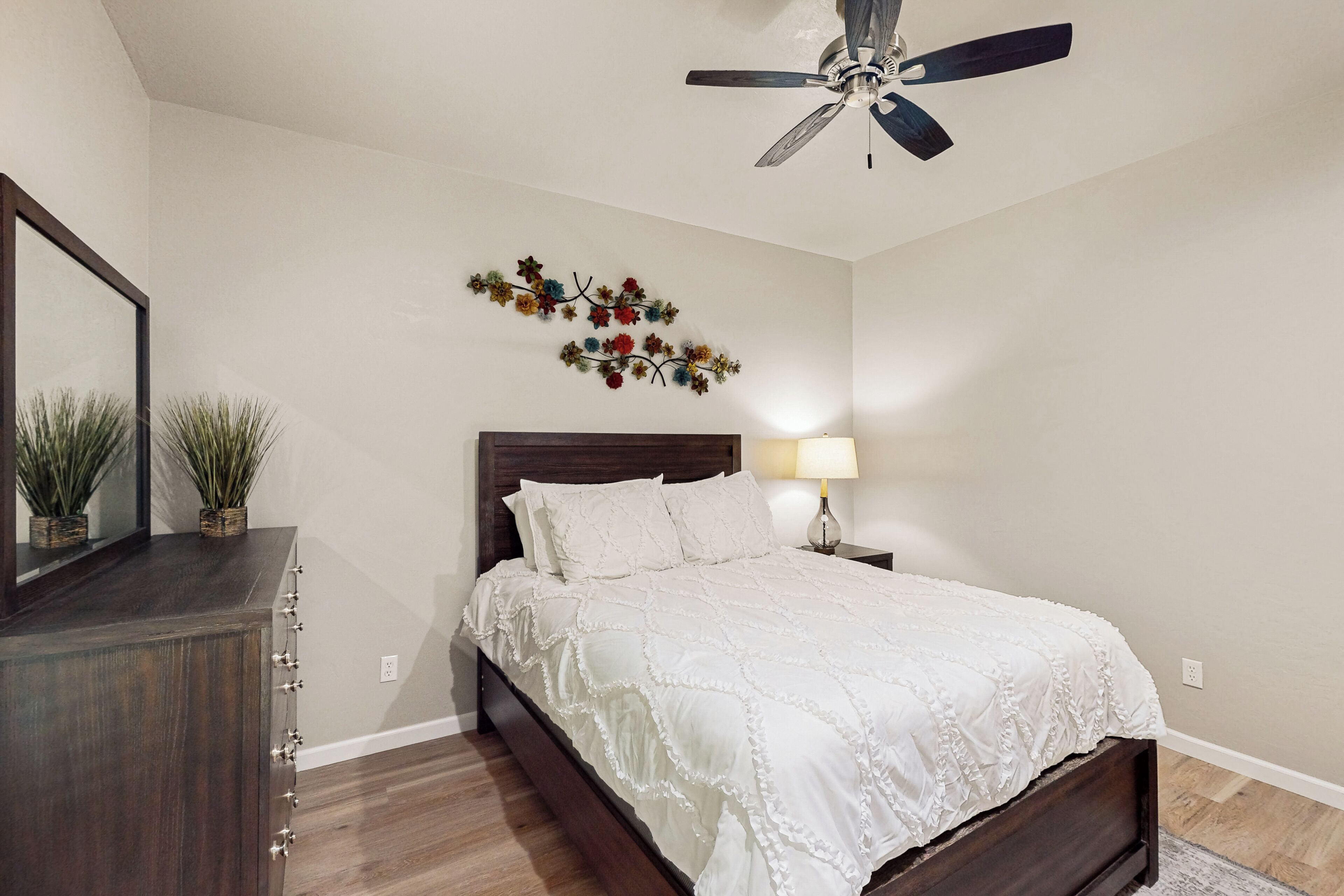 Un dormitorio con una cama de madera, un ventilador de techo, un adorno floral en la pared, una lámpara de noche y un espejo.
