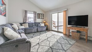 Un salon avec un canapé d’angle gris, un tapis à motifs, un téléviseur posé sur une table en bois et une grande fenêtre avec des rideaux.