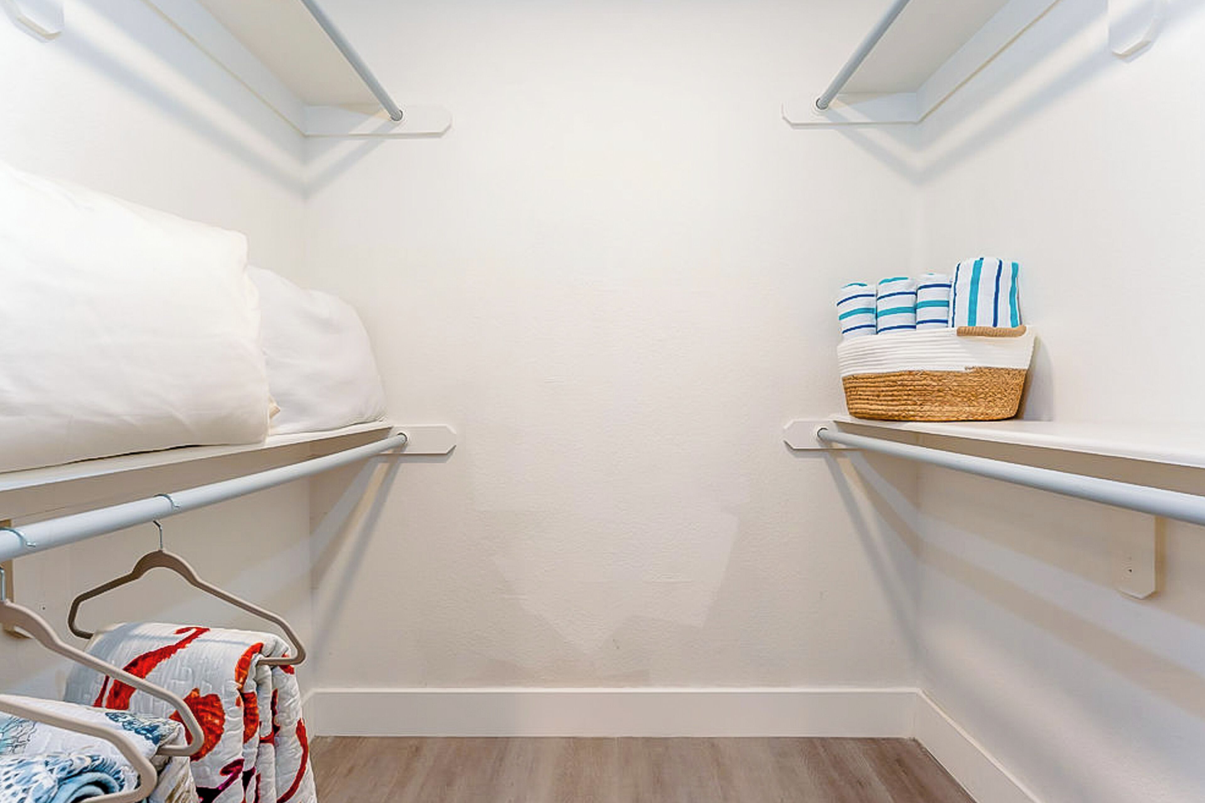 A walk-in closet with white walls and wooden flooring. It features two metal shelves on the left and right, holding folded towels and a basket.