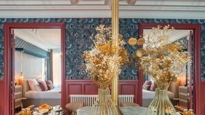 Ein Hotelzimmer mit einem Marmortisch, zwei goldenen Vasen mit Trockenblumen, einem spiegel mit rotem Rahmen und einer Wand mit Muster.