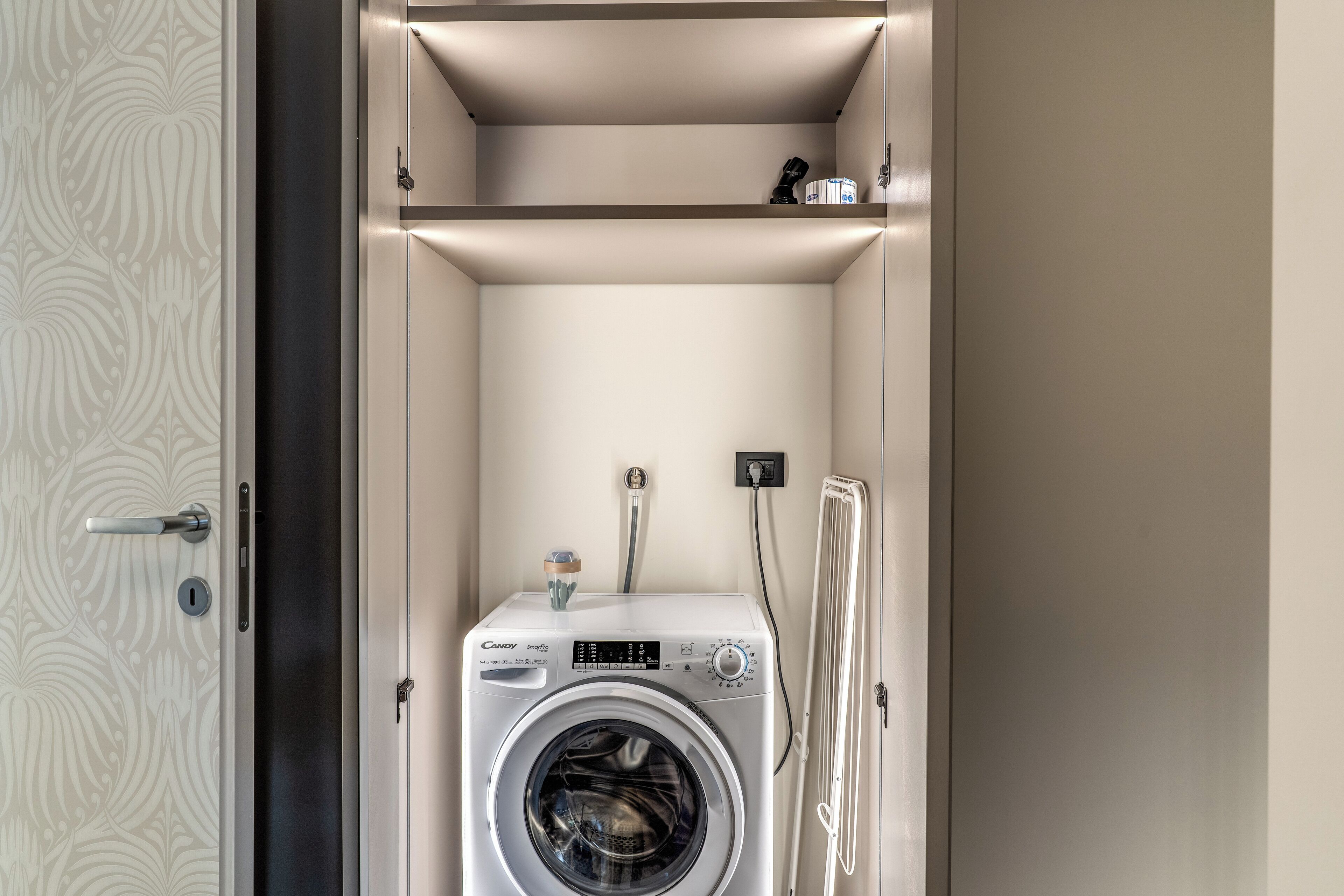 A compact, modern laundry room with a built-in washer and dryer, a small shelf above the dryer, and a door with a decorative pattern.