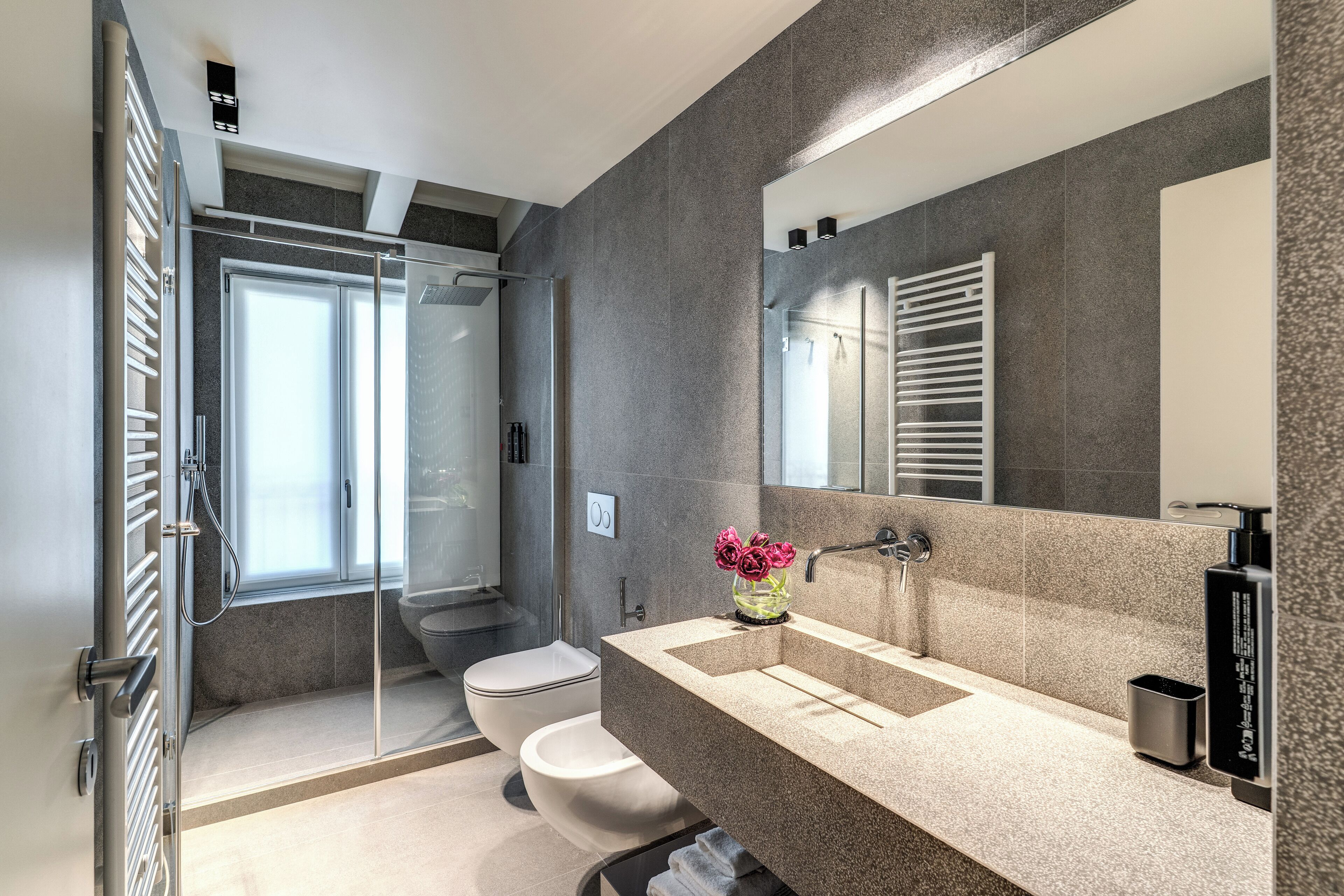 Een moderne badkamer met een douchecabine van glas, een toilet, een wastafel met spiegel en een vaas met bloemen.
