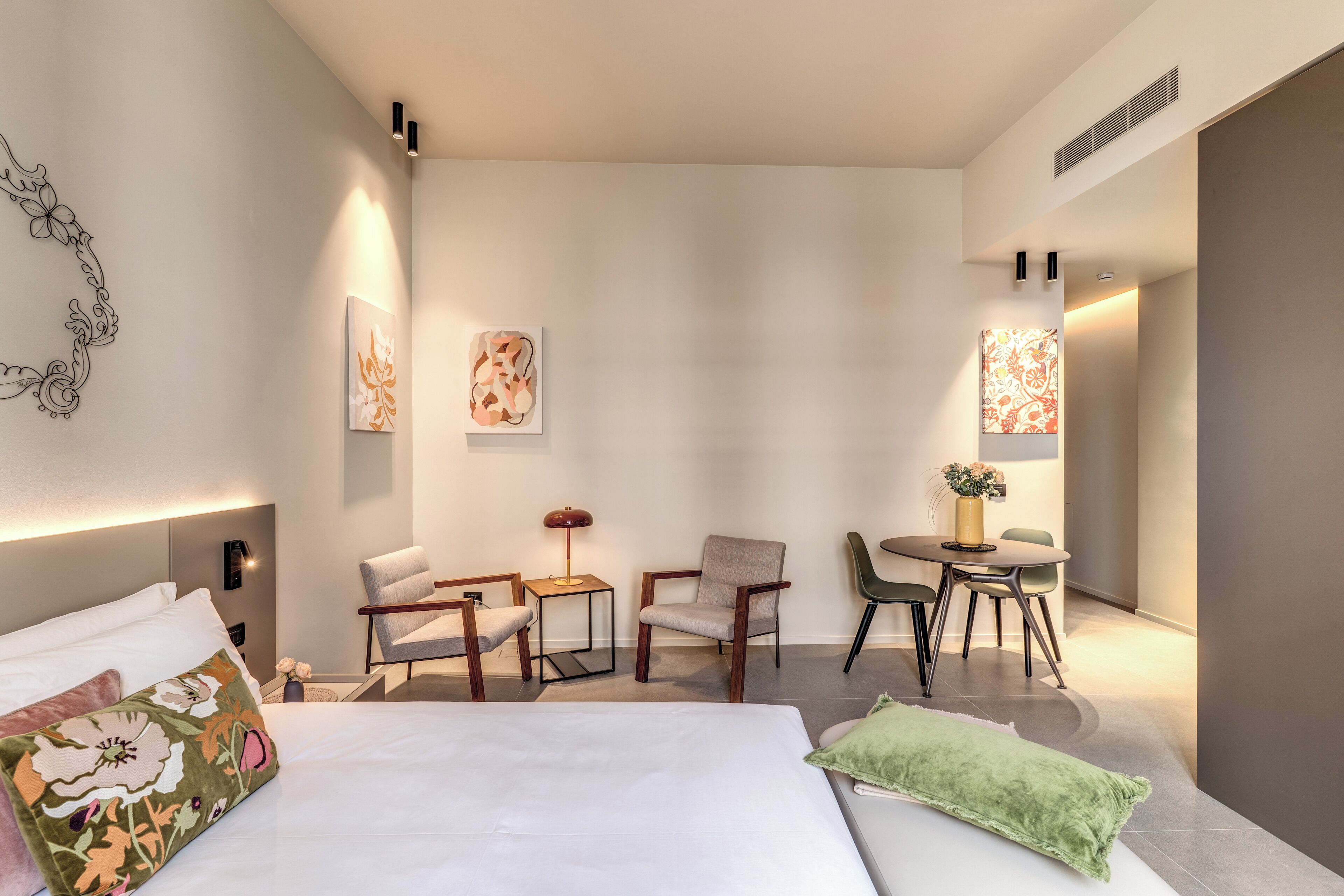 Une chambre d’hôtel moderne, dotée d’un grand lit, de deux fauteuils, d’une petite table et d’un bureau avec des chaises. Des lampes murales et des œuvres d’art ornent les murs.