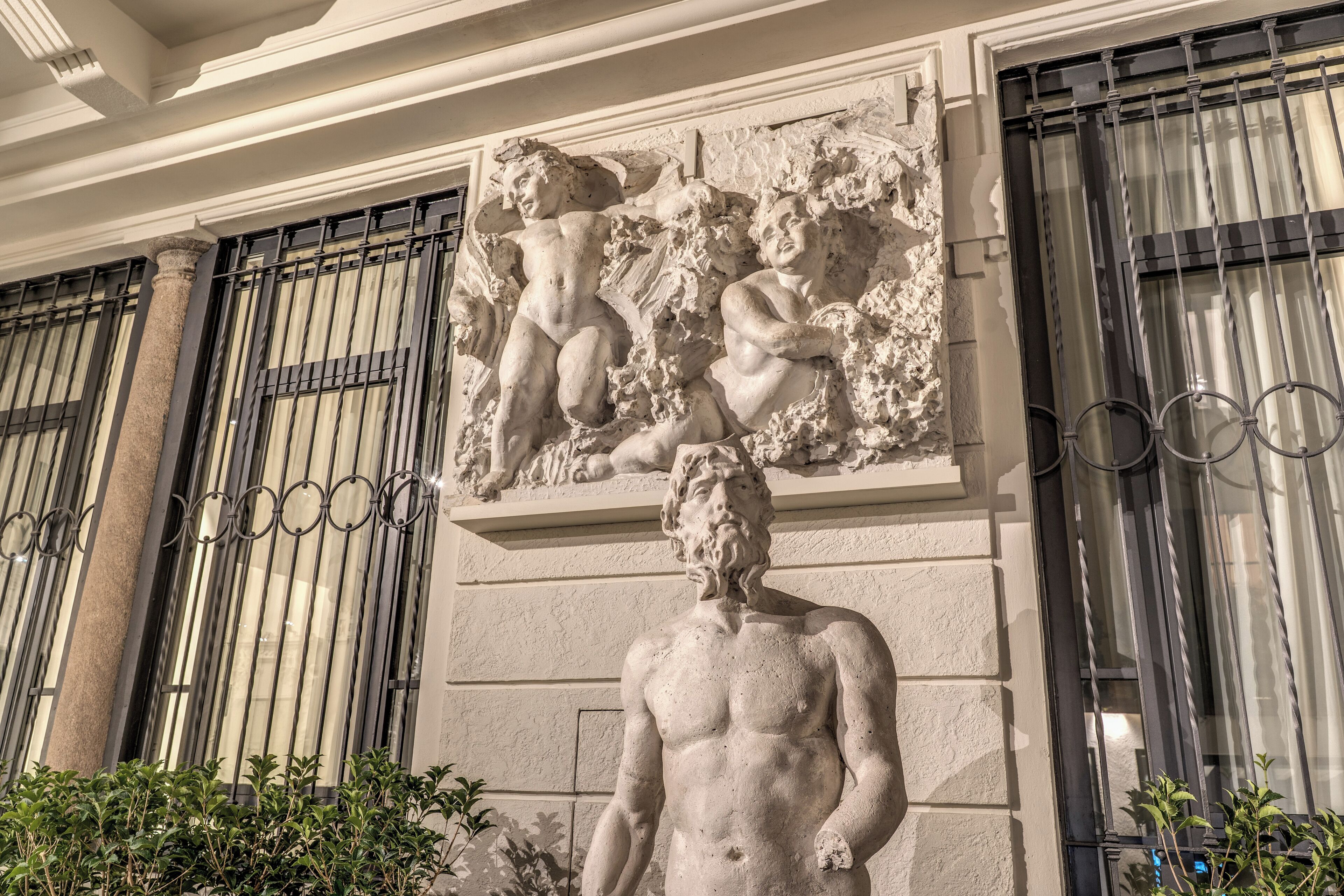 Escultura clàssica a la façana d'un edifici, flanquejada per grans finestres amb reixes metàl·liques.
