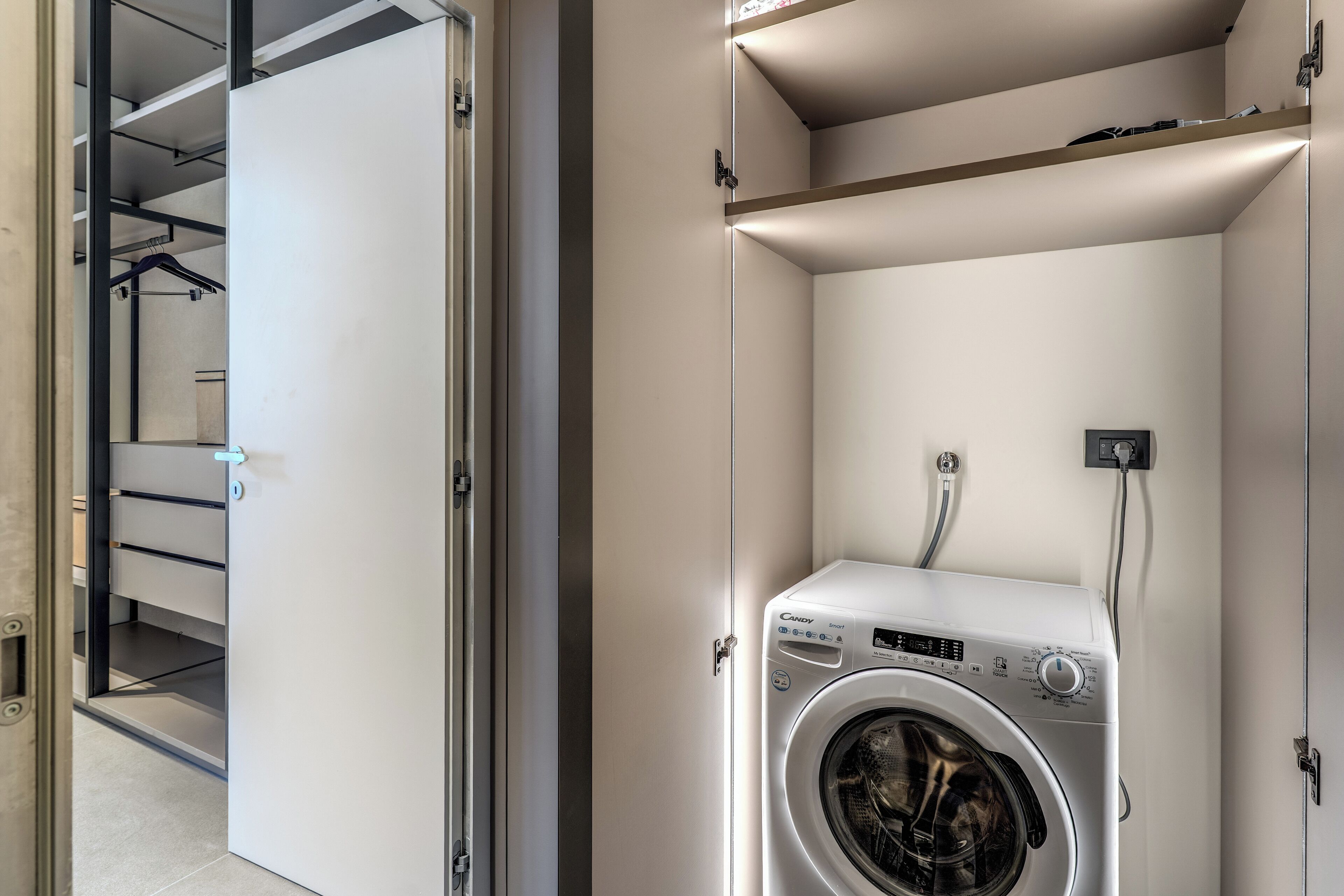 A laundry room with a front-loading washing machine, a shelf above it, and a door leading to a hallway with a coat rack.