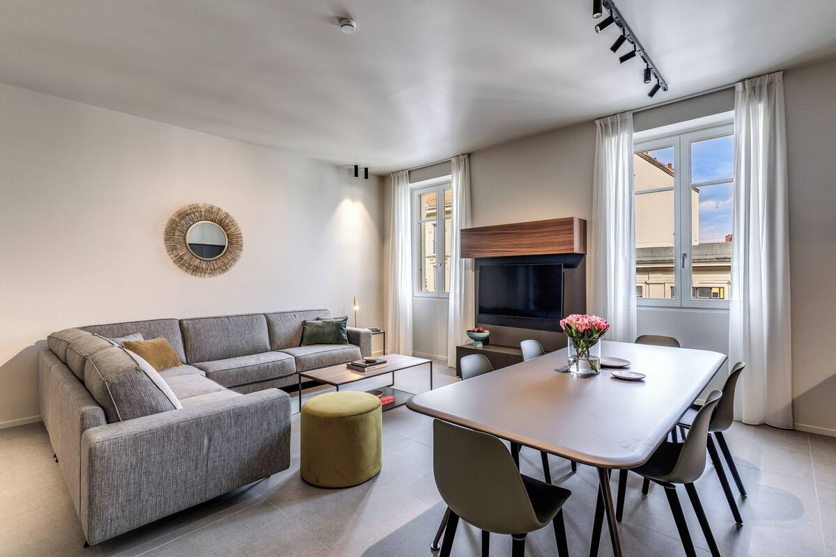 A modern living room with a grey sofa, a dining table with chairs, a round ottoman, a flat-screen TV, and a large window with curtains.